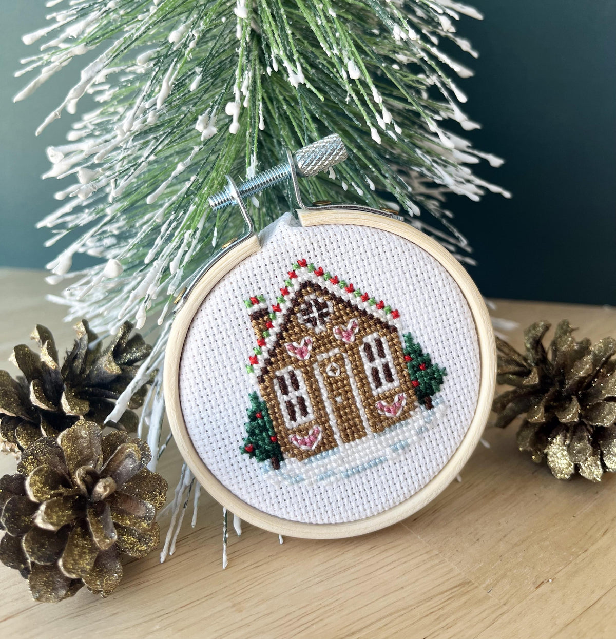 Gingerbread House cross stitch in a small wooden hoop beside a snowy Christmas tree and pine cones