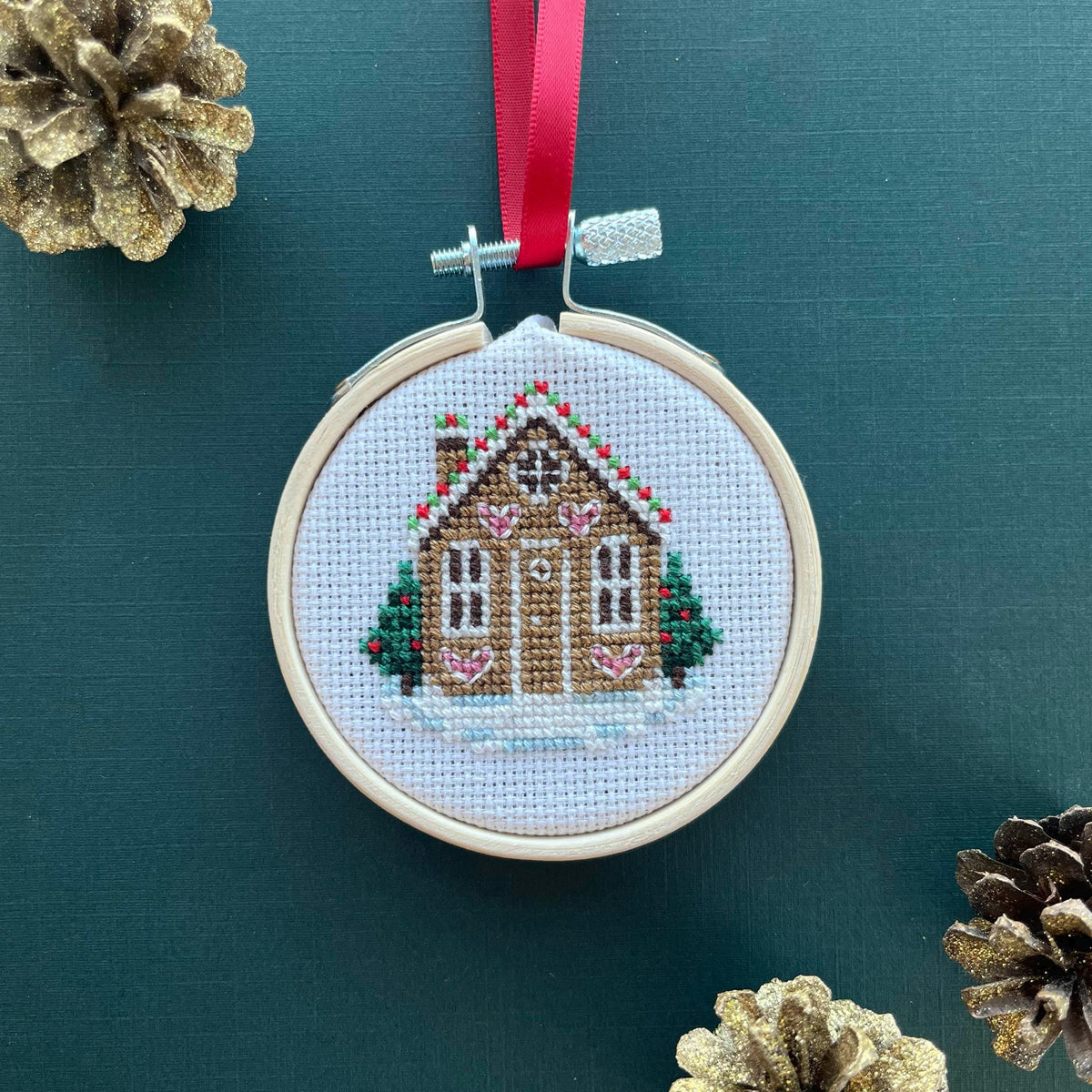 Gingerbread House cross stitch ornament hanging from a red ribbon on a dark green background with gold pine cones